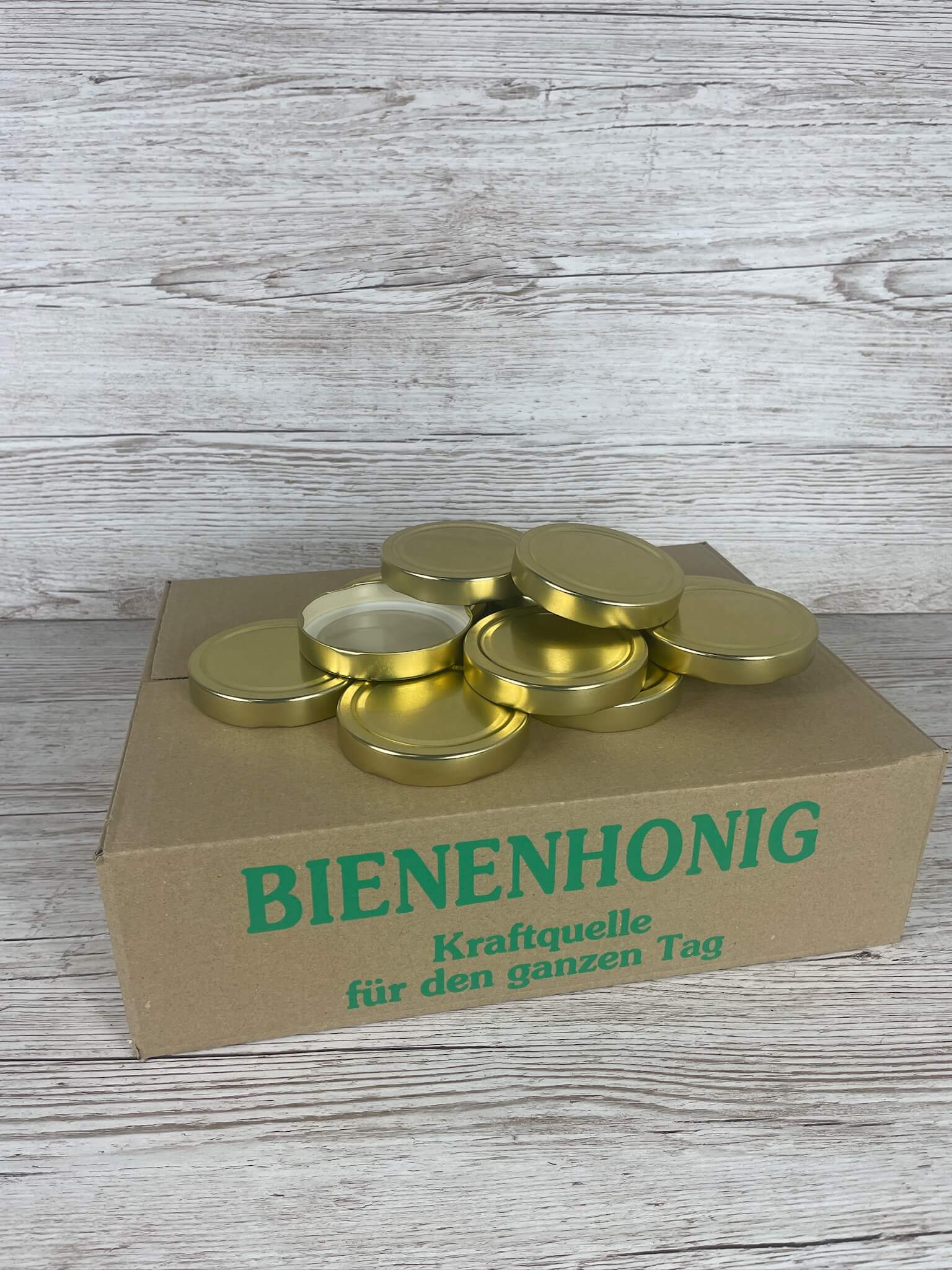 Bienenhonig-Versandkarton für 415ml Gläser mit einem darauf liegenden Stapel goldener Deep-Deckel