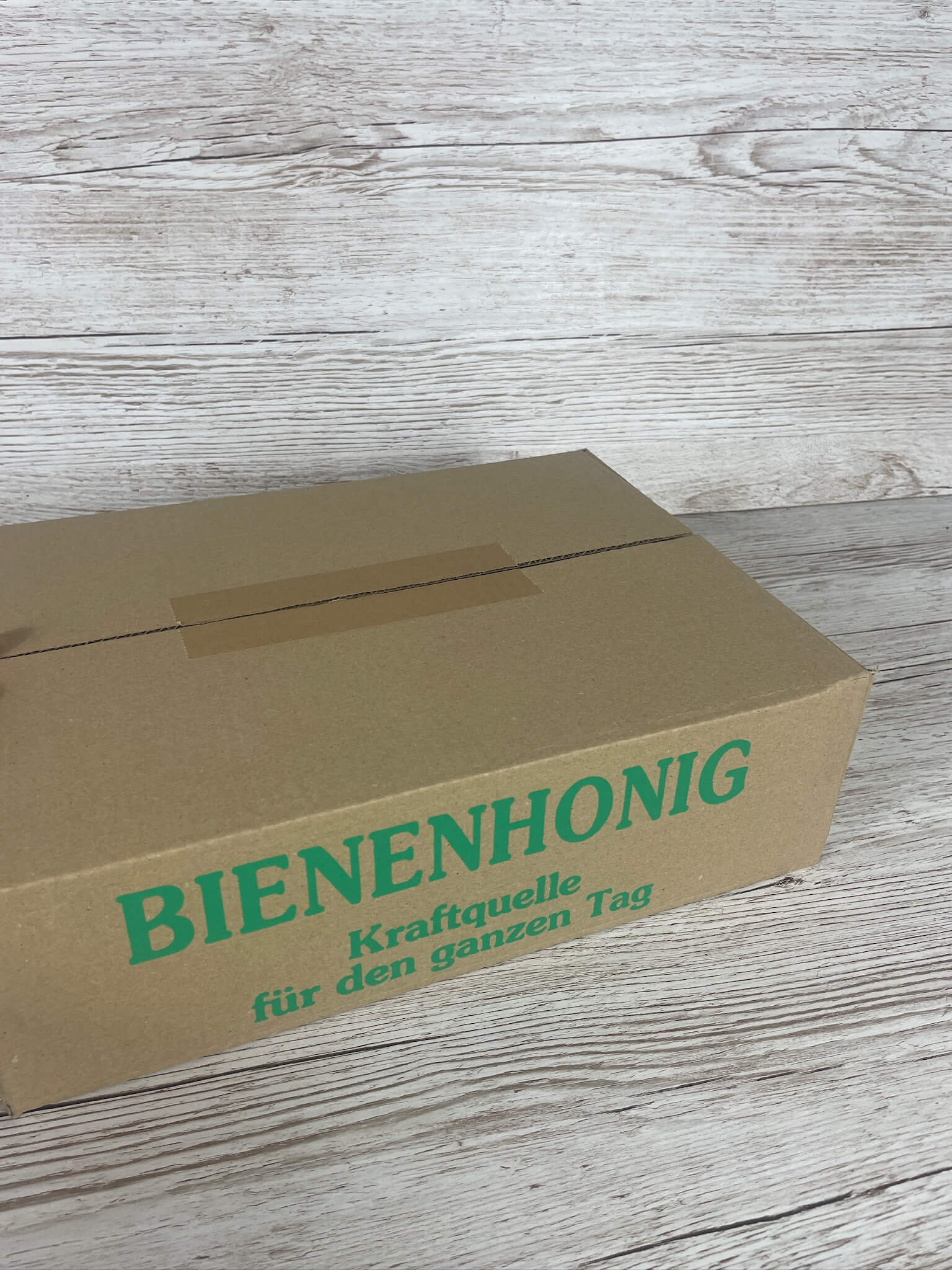 Geschlossener brauner Versandkarton für Honiggläser mit grünem Aufdruck Bienenhonig Kraftquelle für den ganzen Tag