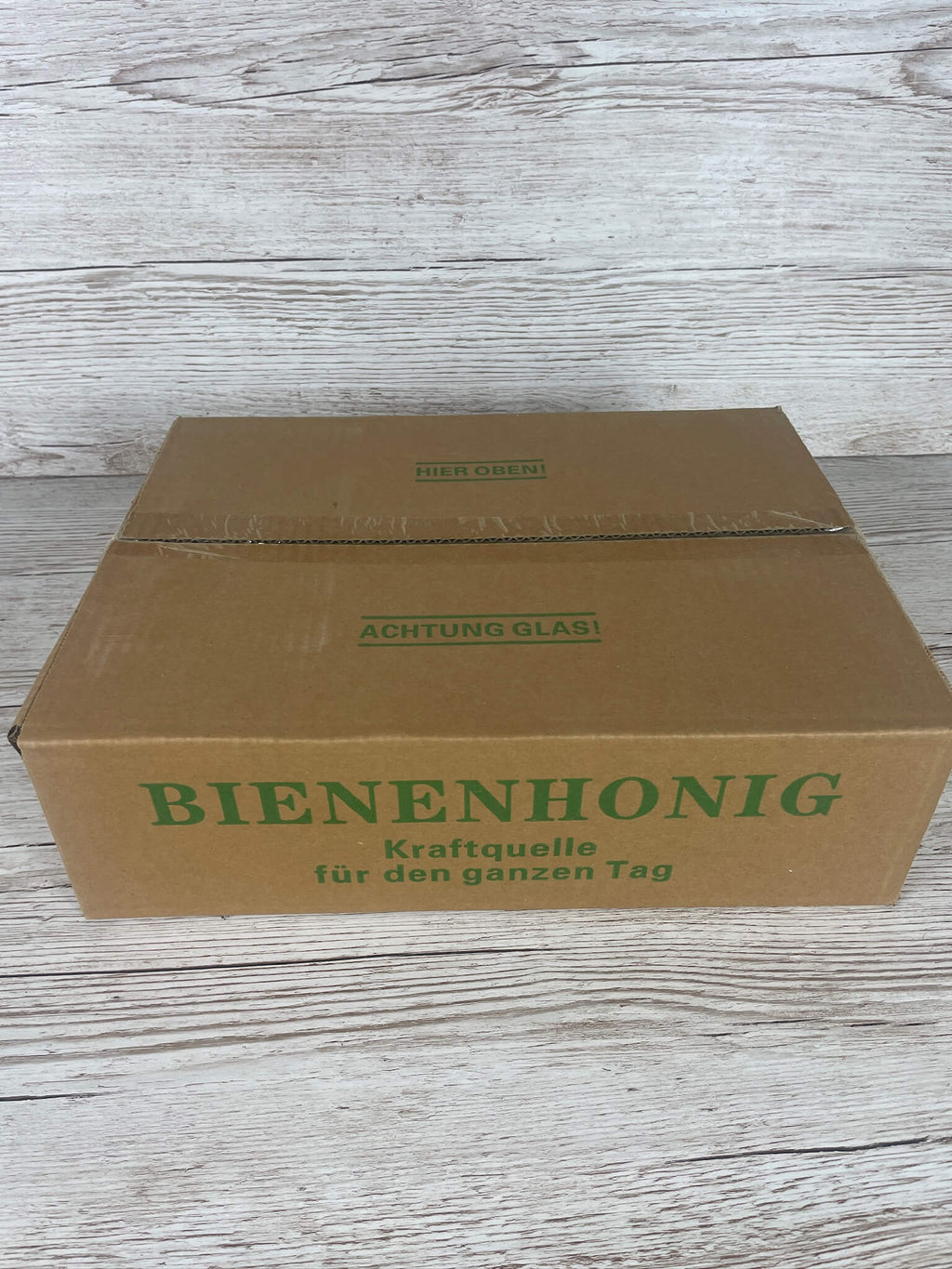 Geschlossener brauner Versandkarton für Honiggläser mit grünem Aufdruck Bienenhonig Kraftquelle für den ganzen Tag und dem Hinweis ACHTUNG GLAS!
