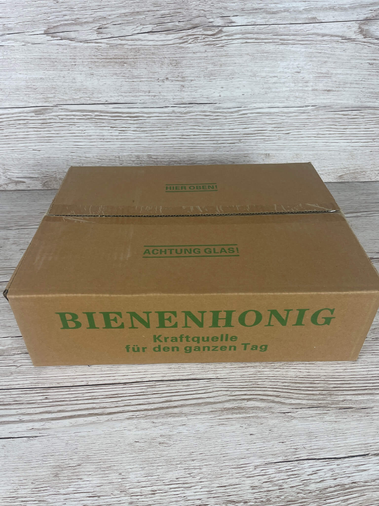 Geschlossener brauner Versandkarton für Honiggläser mit grünem Aufdruck Bienenhonig Kraftquelle für den ganzen Tag und dem Hinweis ACHTUNG GLAS!